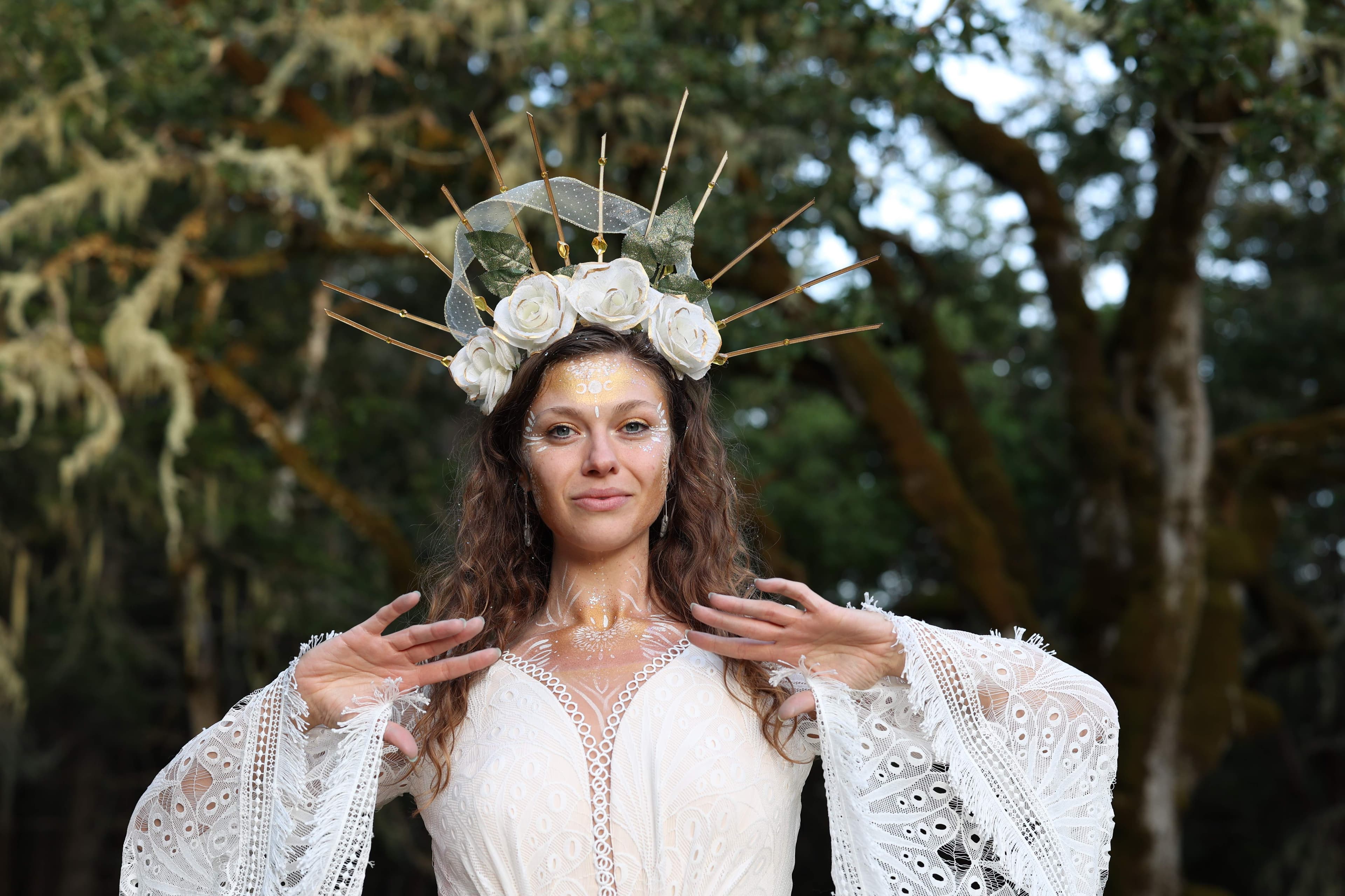 Margo Free in a gold sun crown and white lace, with gold body art, standing in a mossy forest